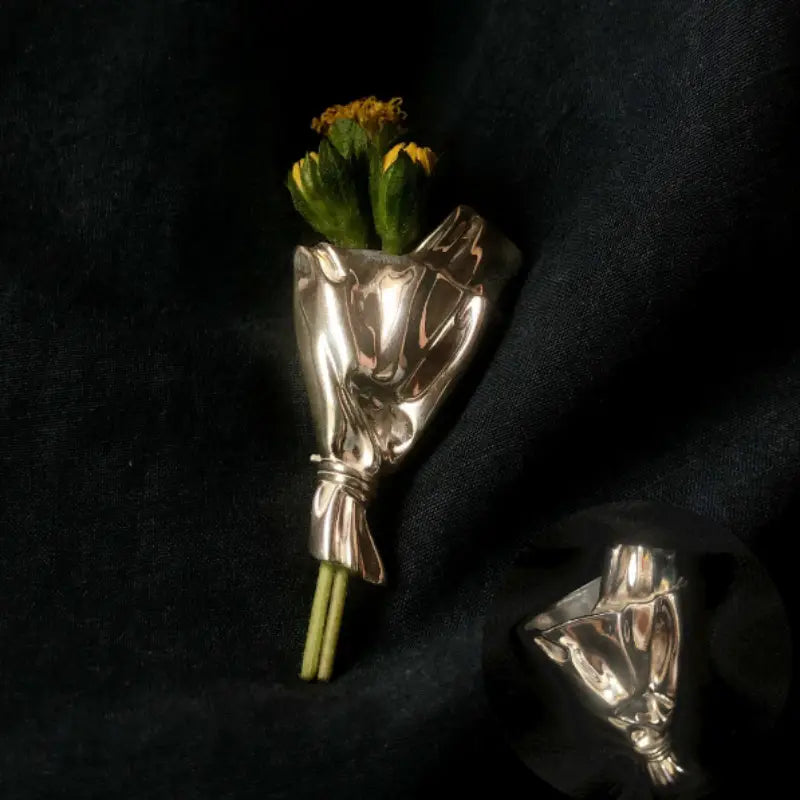 Eine silbermetallische Brosche, gestaltet als Hand, die einen kleinen Strauß gelber Blumen hält, umwickelt mit zartem Papier, zur Verwendung als kreatives Corsage-Zubehör.