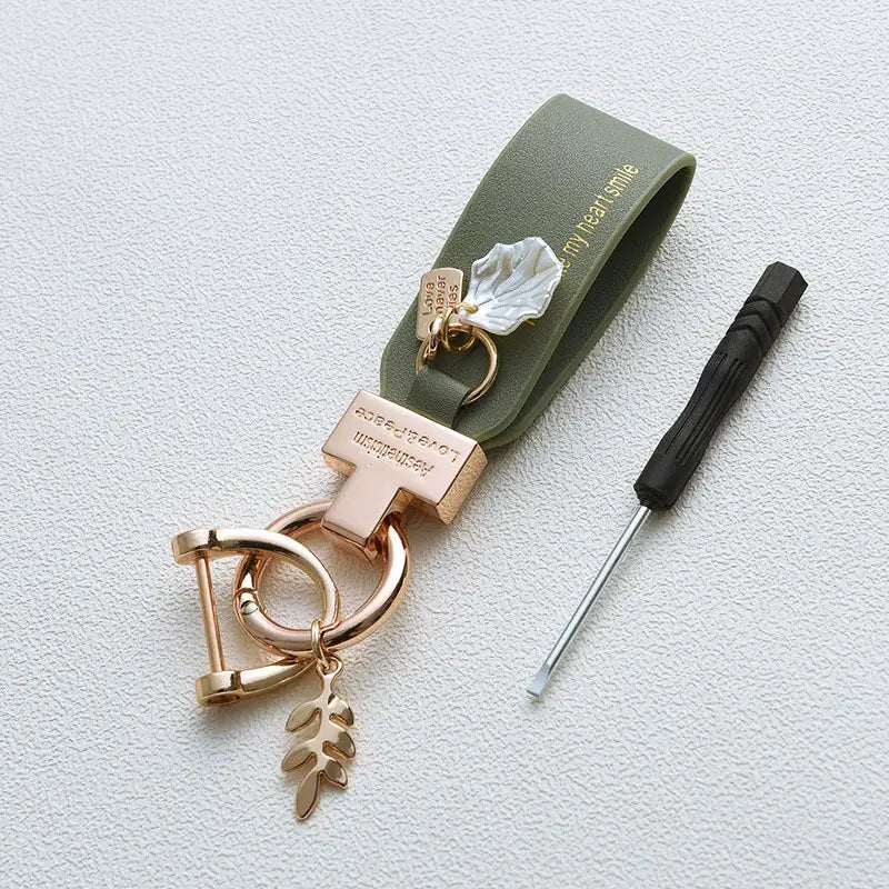 Llavero de piel verde oliva con herrajes metálicos de tono dorado rosado y colgante en forma de concha blanca, diseñado como un llavero antirrobo estilo francés para automóvil.