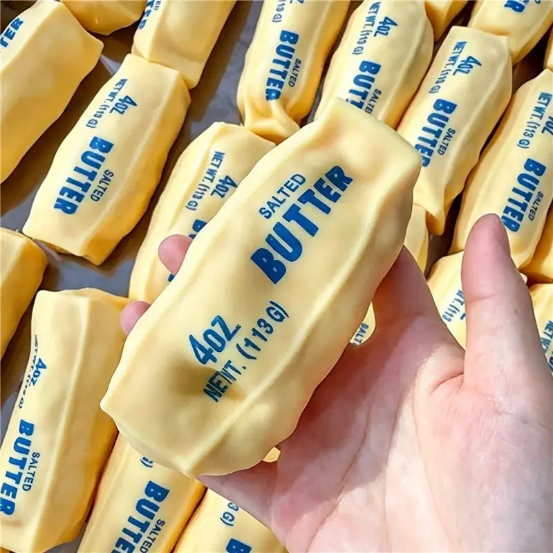 Eine Hand hält einen gelben rechteckigen Butterstick als ASMR-Sensory-Squishy-Spielzeug mit blau bedrucktem Text, der Gewicht und Inhalt angibt.