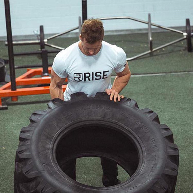 Ein schwarzer Gummireifen mit tiefen Profilen und eingeprägtem Text an der Seitenwand, der zusammen mit einem Herren-Workout-T-Shirt mit Gold-Rise-Logo dargestellt wird.