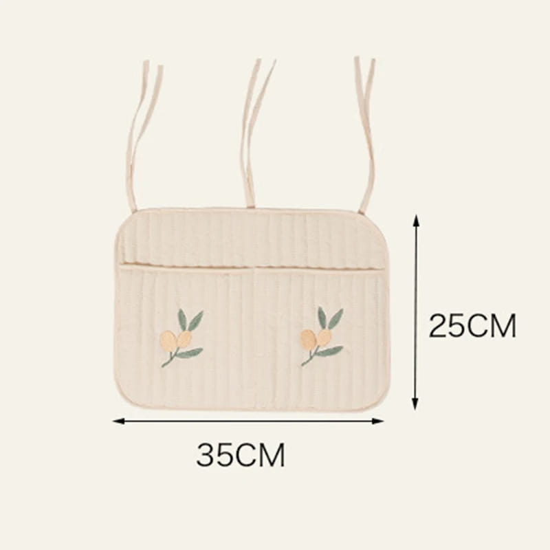 ベージュ色のキルティング製ベビーカー用オーガナイザー袋で、刺繍のモチーフ（桃）と、ベビーカーでの使用に便利な布製の細いストラップが2本付いています。