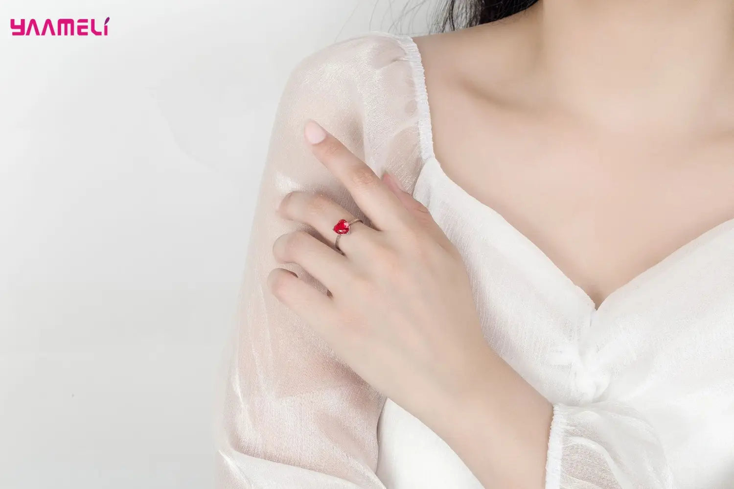 A delicate red heart-shaped sterling silver ring with a shiny metallic band worn on the model’s finger from the sterling silver heart jewelry set zircon huggies.