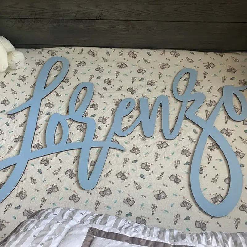 Hellblauer Holz-Aufschnitt mit personalisiertem Babynamenschild, Buchstabierung „joffenzo“ in geschwungener Kurrentschrift, für die Wanddekoration im Kinderzimmer.