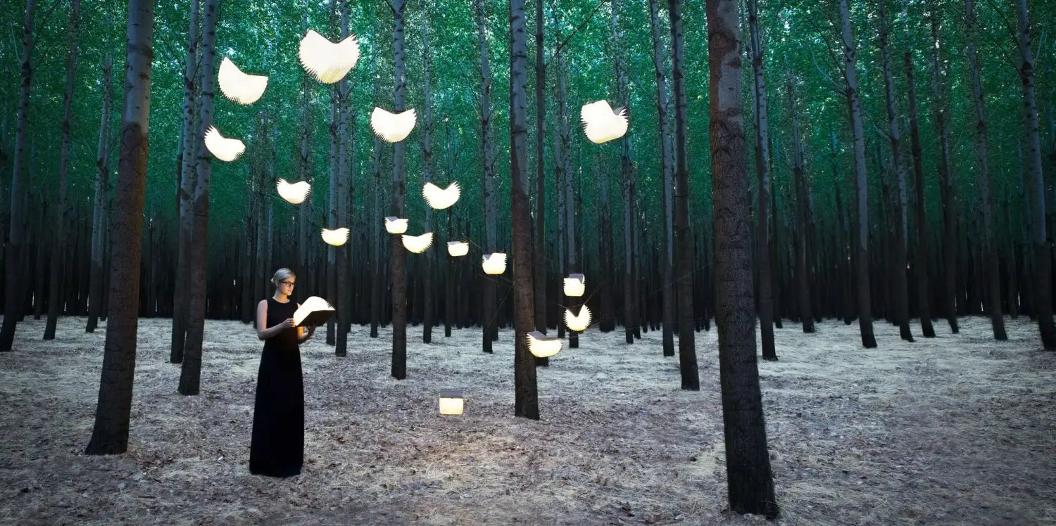 Una mujer con un vestido negro se encuentra bajo linternas de luz LED plegables con forma de libro que emiten un brillo, suspendidas entre árboles altos en un entorno al aire libre.