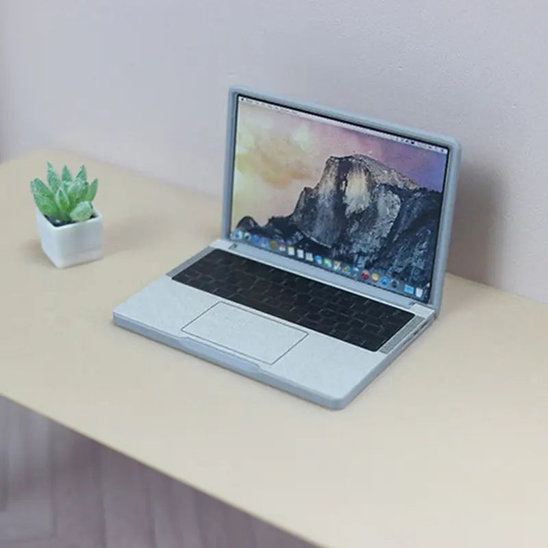 Silberne MacBook-Miniatur-Laptop-Puppenstuben-Zubehör mit schwarzer Tastatur und Trackpad, das auf dem Bildschirm Berg-Wallpaper anzeigt.
