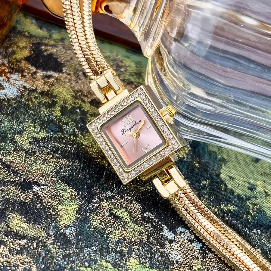 Gold diamond square watch with a gold-toned case, pink dial, roman numerals, diamond bezel, and an ornate snake-like fringe strap.