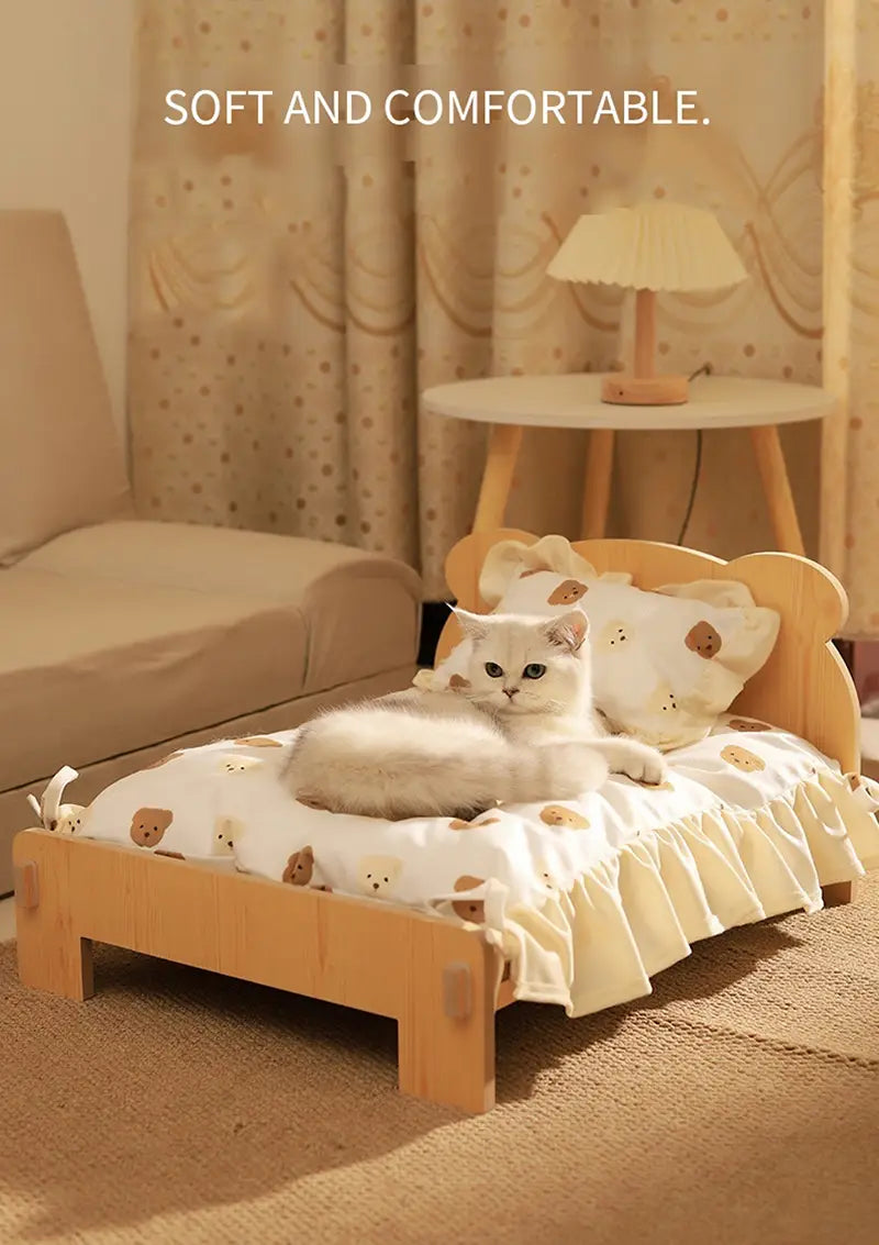 A cozy wooden cat bed featuring a light wood frame and a soft white fabric cover with small brown bear faces and ruffled edges.