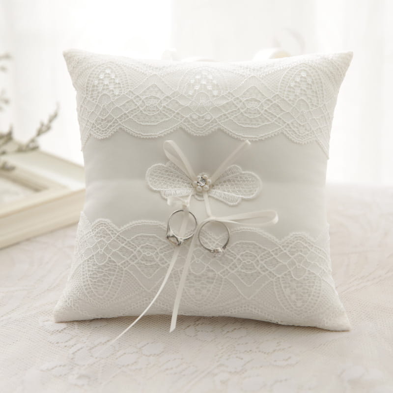 Almohada blanca para anillos de boda de encaje con un delicado lazo y un realce de cristal, diseñada como almohadilla portadora de arras.