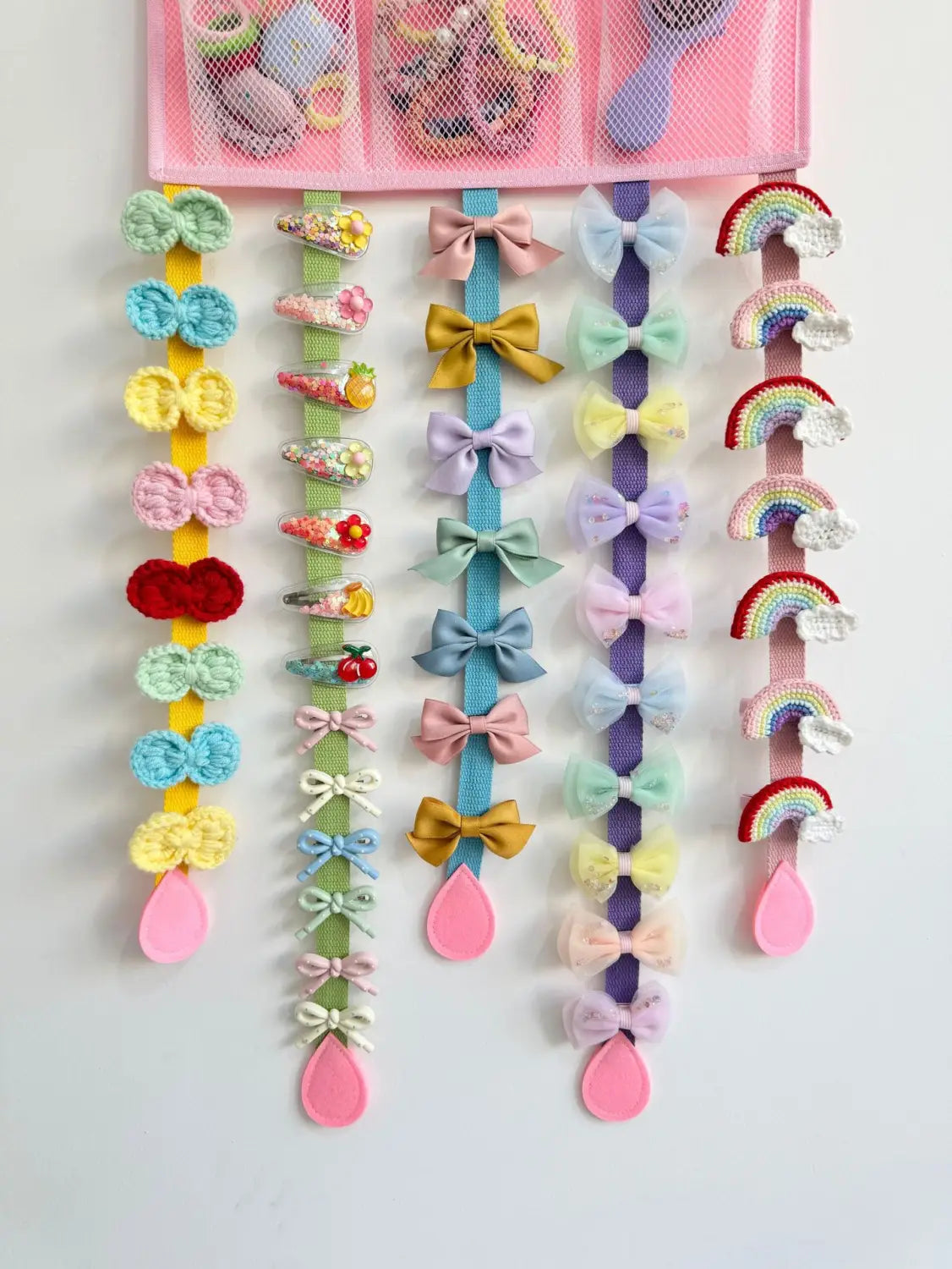 Lazo tejido a crochet en tonos pastel y clips de cabello con temática de arcoíris dispuestos sobre bandas elásticas de colores y mostrados en un organizador colgante para accesorios de cabello con temática de arcoíris.