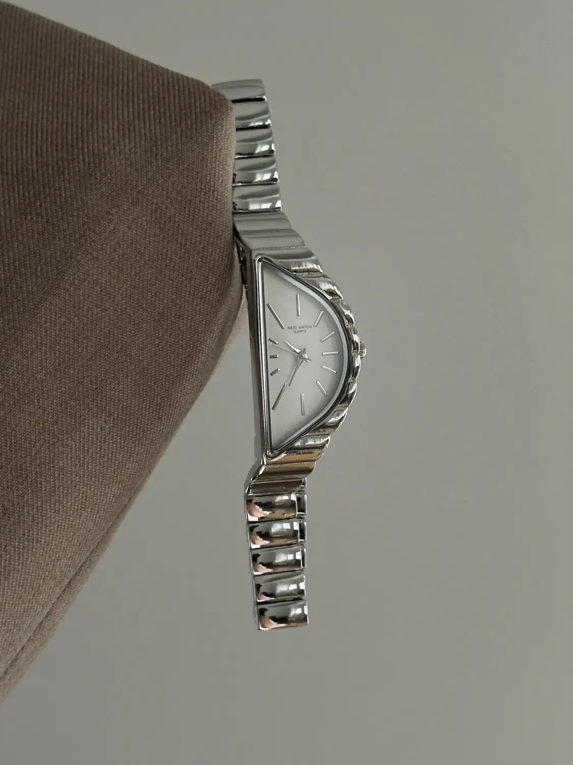 A sleek silver-toned geometric triangle quartz watch made of stainless steel with a distinctive triangular face and ridged bracelet featuring minimalist hour markers on its white dial.
