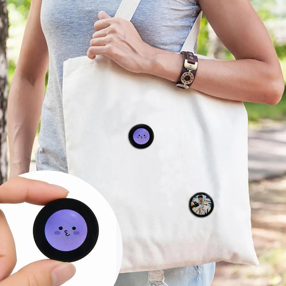 White canvas tote bag adorned with two circular black-rimmed stickers featuring a purple cartoon face and a photo of a person promoting a mini gadget smart desktop companion with data cable.