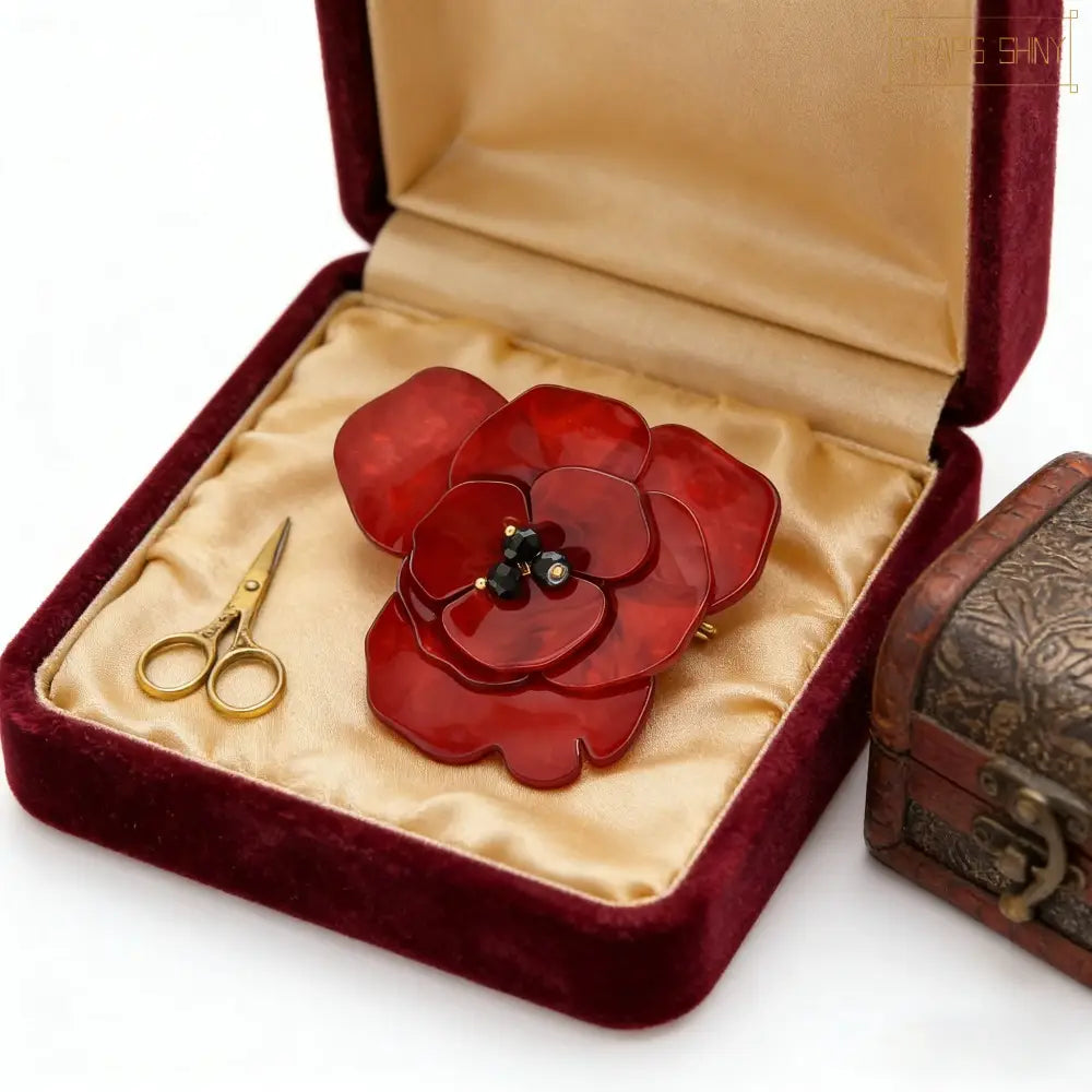 Une broche en émail rouge laqué, en forme de coquelicot, avec des pétales superposés et un accent de centre sombre, présentée dans une élégante boîte en velours.