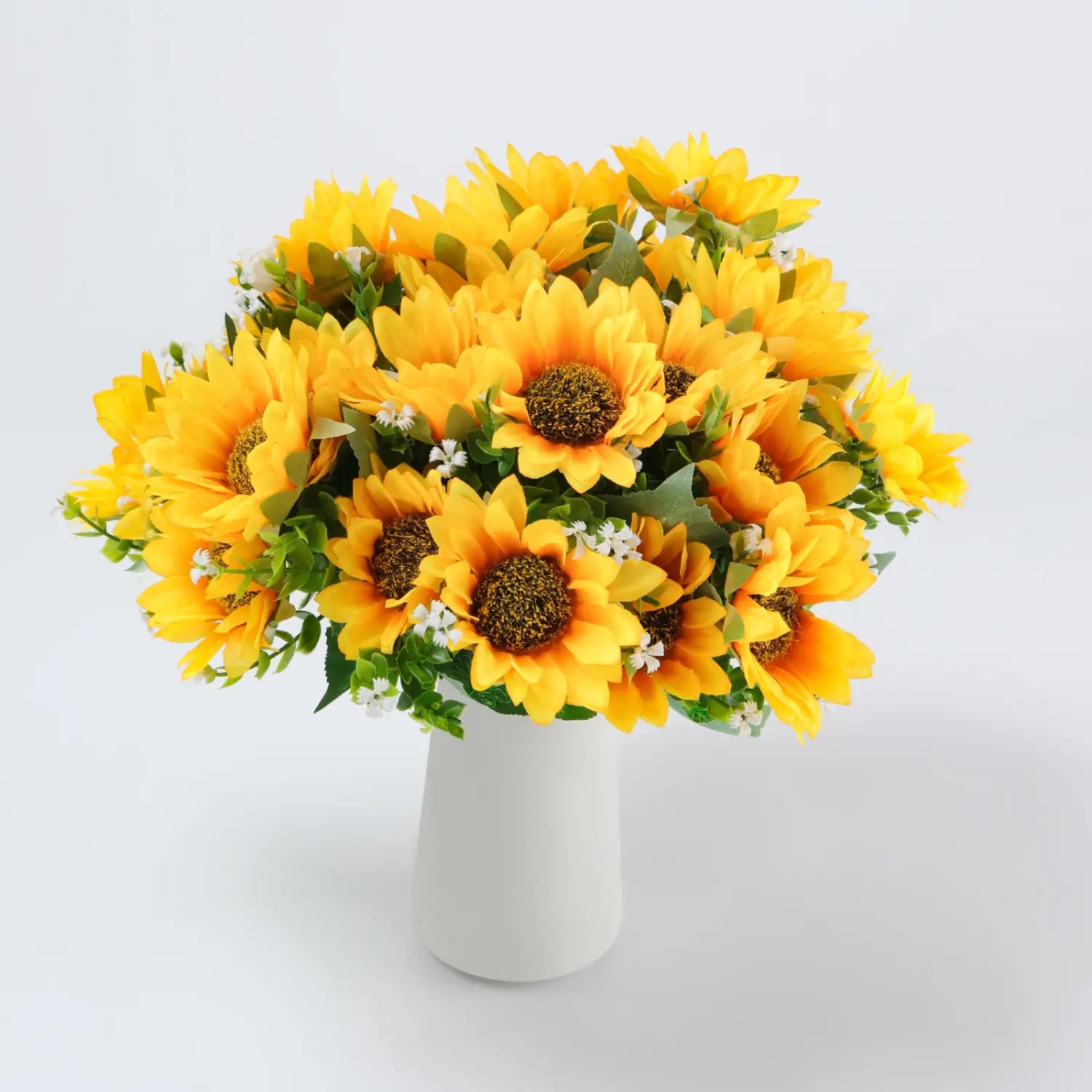 Ein lebendiger Kunstblumenstrauß aus Sonnenblumen mit leuchtend gelben Blütenblättern, dunkelbraunen Zentren, kleinen weißen Blüten und grünen Blättern, angeordnet in einer schlichten weißen Keramikvase.