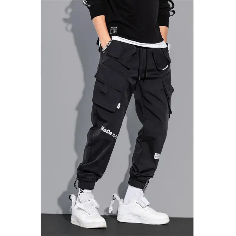 Black mens tactical cargo pants with multiple pockets, white branding on the leg, and a drawstring waistband for streetwear use.