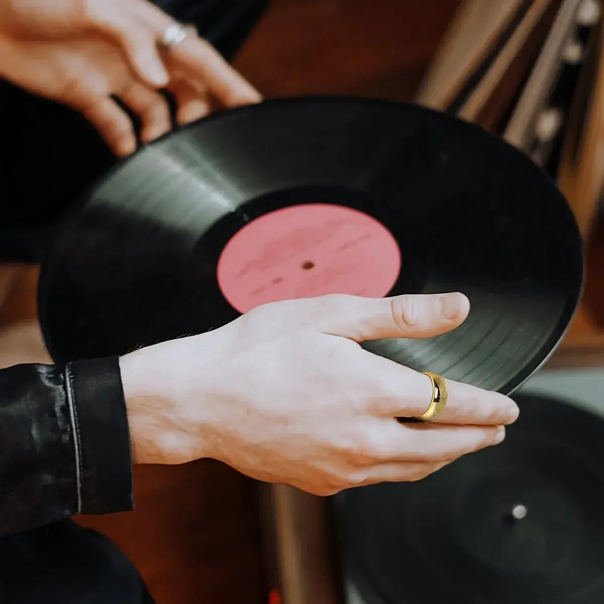 hands holding a black vinyl record with a pink center label alongside a stainless steel elvish script golden wedding band ring