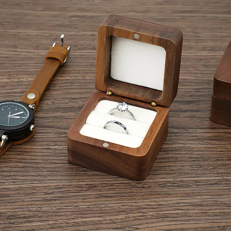 Une boîte à bague en bois brun foncé avec couvercle à charnières et inserts en mousse blanche tenant une bague d’engagement en diamant et une alliance unie pour un usage lors de la proposition et de l’engagement.