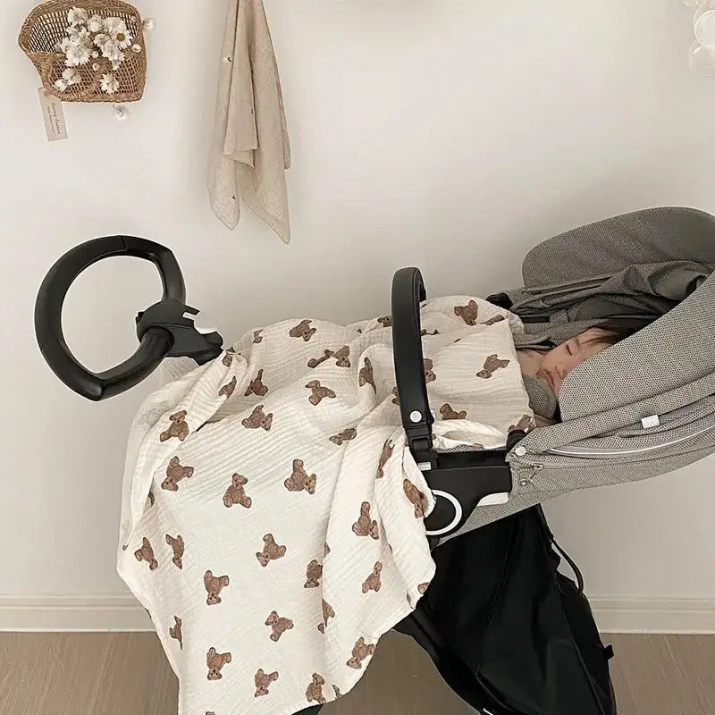 ein weiches Baby-Badehandtuch und eine Wickeldecke aus Muslin in sanftem Off-White mit Bärenmotiv in Braun; das Muster aus braunen Teddybären fällt über einen Kinderwagen