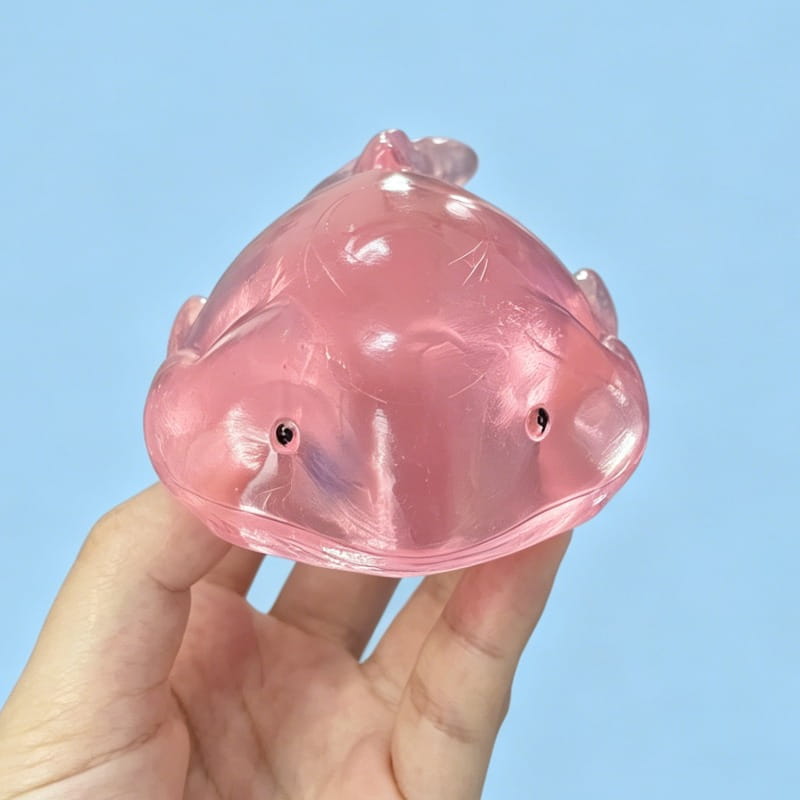 un jouet anti-stress translucide rose en forme d’axolotl, avec des yeux à perles noires, fabriqué en matériau souple et extensible, inclus dans un coffret de jouets anti-stress à mâcher « baleine » en matériau souple