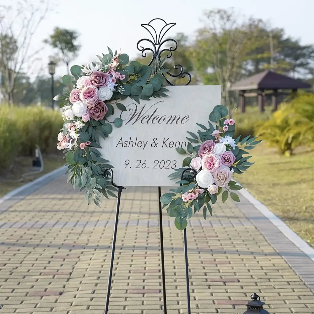 Un letrero decorativo de bienvenida de un juego de flores para arco de boda con rosas artificiales, que presenta rosas artificiales en tonos rosa y blanco y hojas de eucalipto verdes, montadas sobre un soporte negro de hierro forjado