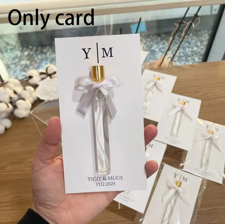un flacon de parfum en verre transparent avec un capuchon doré et un délicat ruban argenté noué autour de son goulot, présenté sur une carte blanche portant le logo y|m, en tant que carte cadeau de parfum de mariage personnalisée, faveur personnalisée