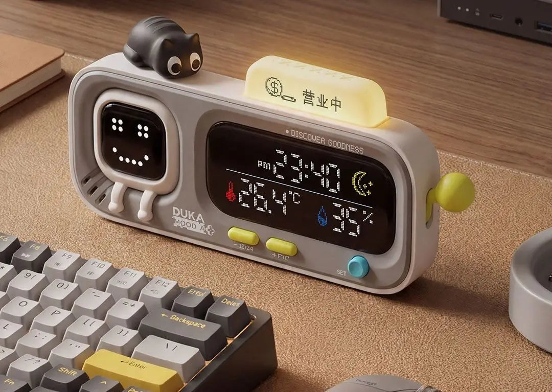 gray duka mooda smart temperature and humidity clock with a smiling face display and a small black cat figurine on top showing temperature and humidity readings