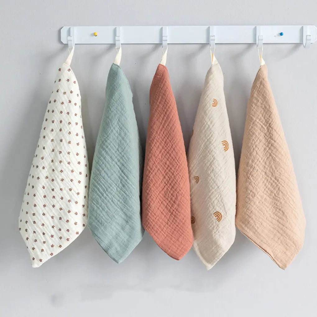Fünf weiche, strukturiere Mulltücher in Pastellfarben, darunter Weiß mit braunen Sternen, Mintgrün, Terrakotta, Creme mit goldenen Regenbögen sowie Beige, ordentlich an einem weißen Regal als Teil eines Bio-Baumwoll-Badetuch- und Mulltuch-Sets aufgehängt.