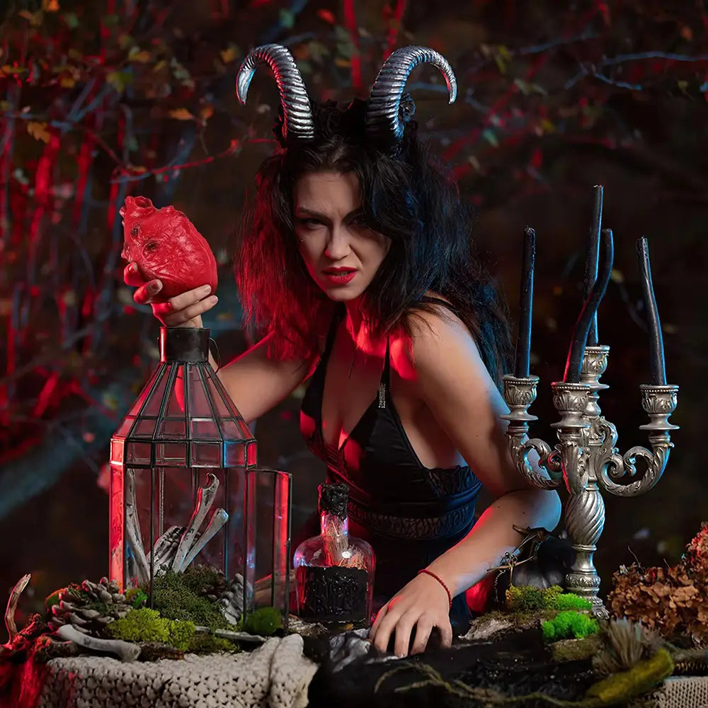 A woman with dark hair and a silver horned headpiece holds a fake bloody human heart Halloween prop made of red textured material.