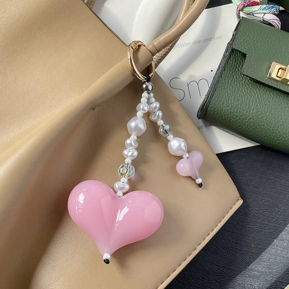 Pendentif pour sac à perles en forme de cœur, comprenant une charm en verre rose en forme de cœur brillante ornée de perles blanches et de cristaux transparents, suspendue à un anneau métallique à finition dorée.