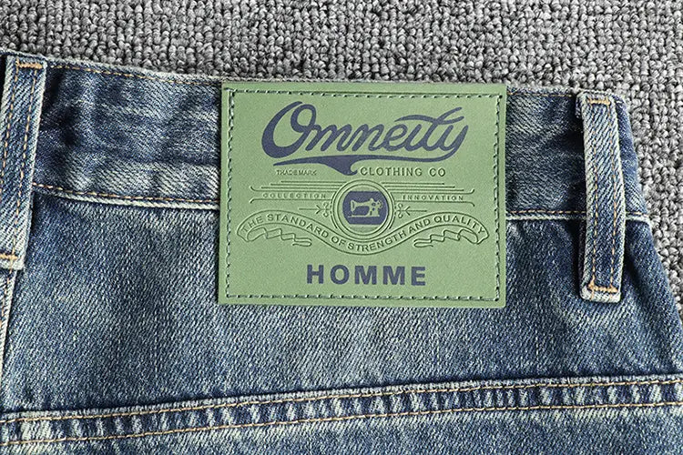 Jeans en denim bleu clair avec une coupe ample brodée pour hommes, comportant un écusson en cuir vert avec le nom de la marque Omneity et le mot Homme imprimé en dessous à l’arrière.