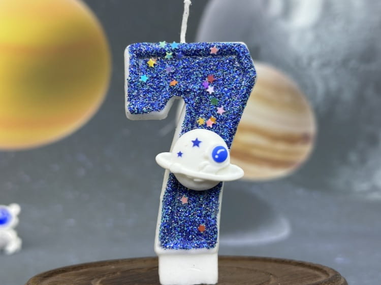 Una vela azul con brillo con el número siete para cumpleaños, con chispas con forma de estrellas de colores y una figura de planeta blanco con ojos azules adherida, diseñada como adorno para pastel de fiesta planetaria.