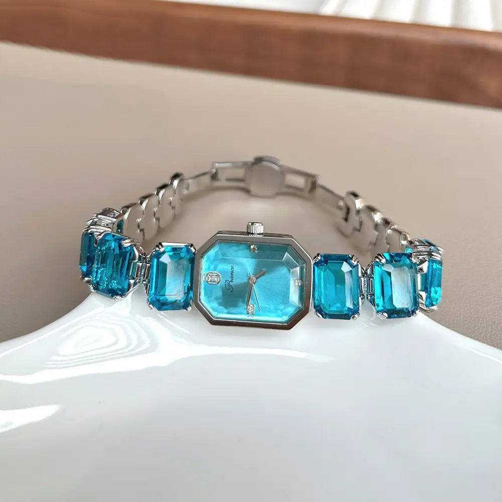 A sleek silver-toned women quartz crystal bracelet watch with a rectangular turquoise face and four matching emerald-cut blue gemstones set along its band.