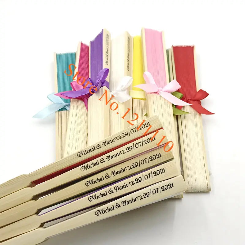 A set of light-colored wooden folding fans made of bamboo with colorful ribbon bows and engraved personalized wedding details as custom wedding hand fans and silk favors.