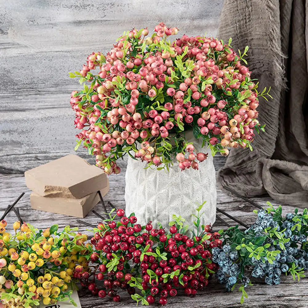 Une branche luxuriante de fleurs de baie artificielles simulées, en mousse, avec des baies rosâtres-rouges et des feuilles vertes, disposées dans un vase en céramique blanche texturée pour la décoration de la maison et des mariages.