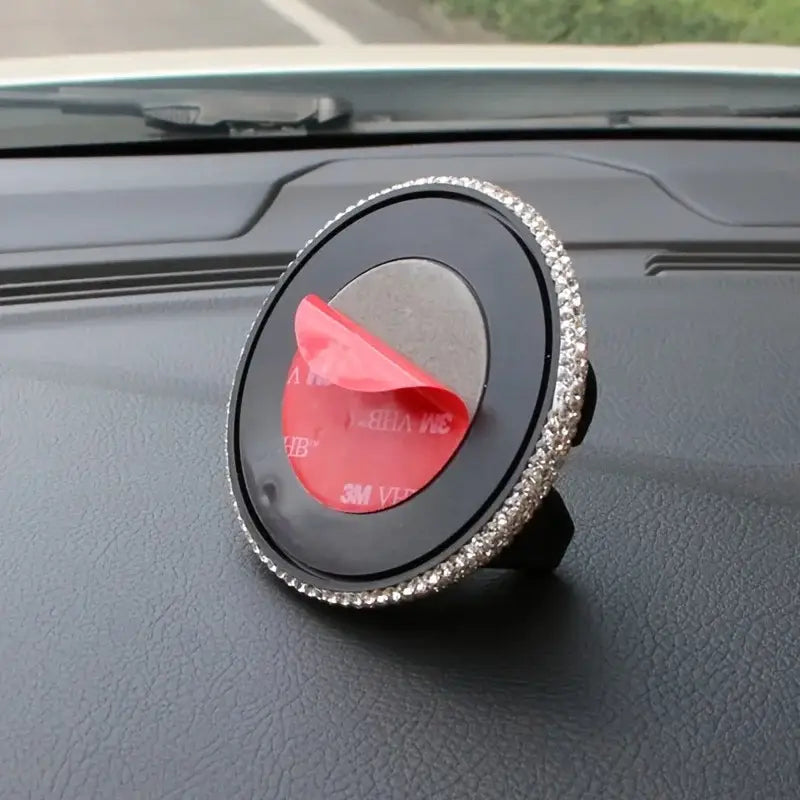 Un soporte circular negro para teléfono de automóvil con ventilación, de pedrería, con un borde de pedrería brillante plateada y adhesivo rojo de 3M, mostrado desde el lado posterior.