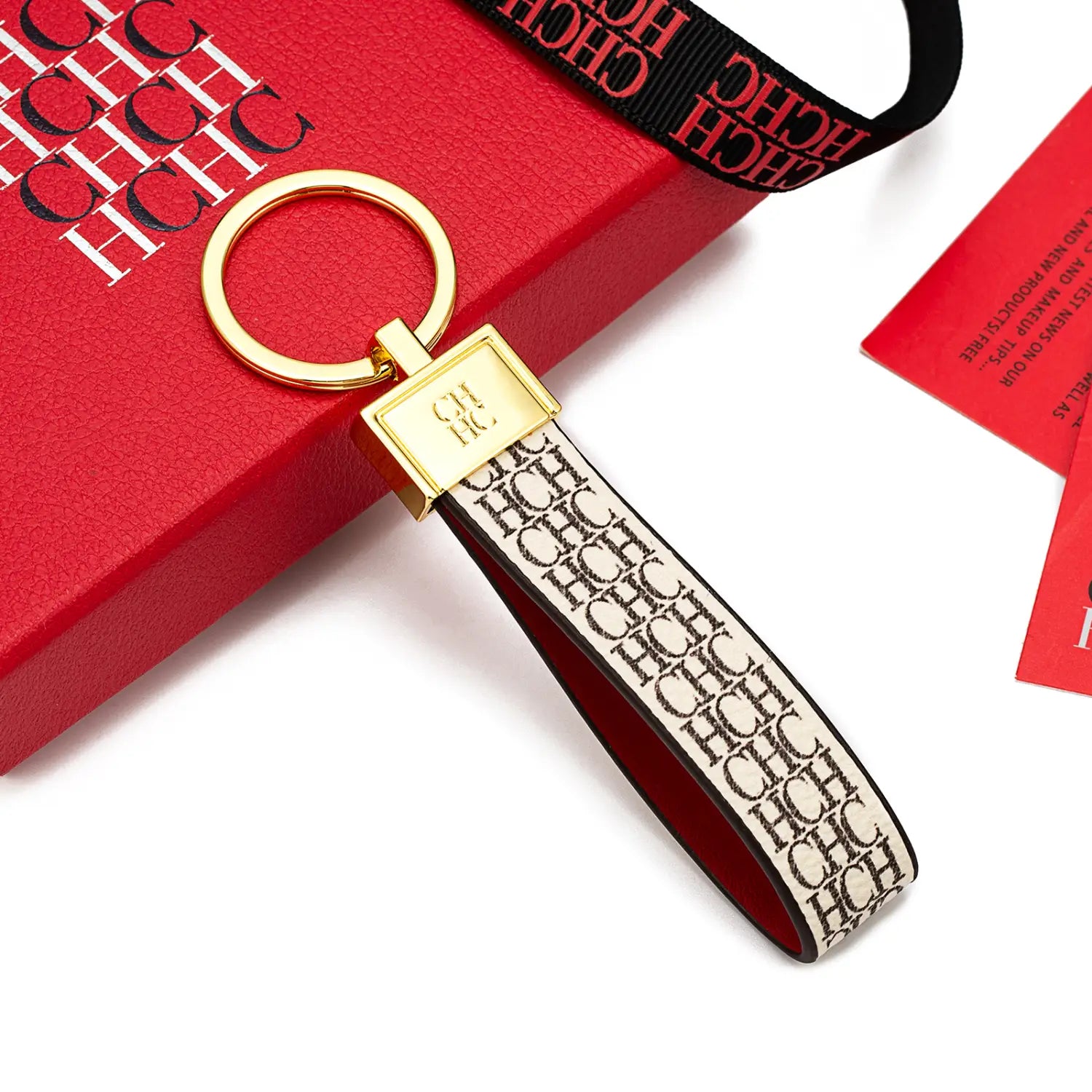 Gold-tone key ring with a rectangular plaque and repeating chic logo on an off-white leather strap leather logo keychain designer fashion accessory gift.