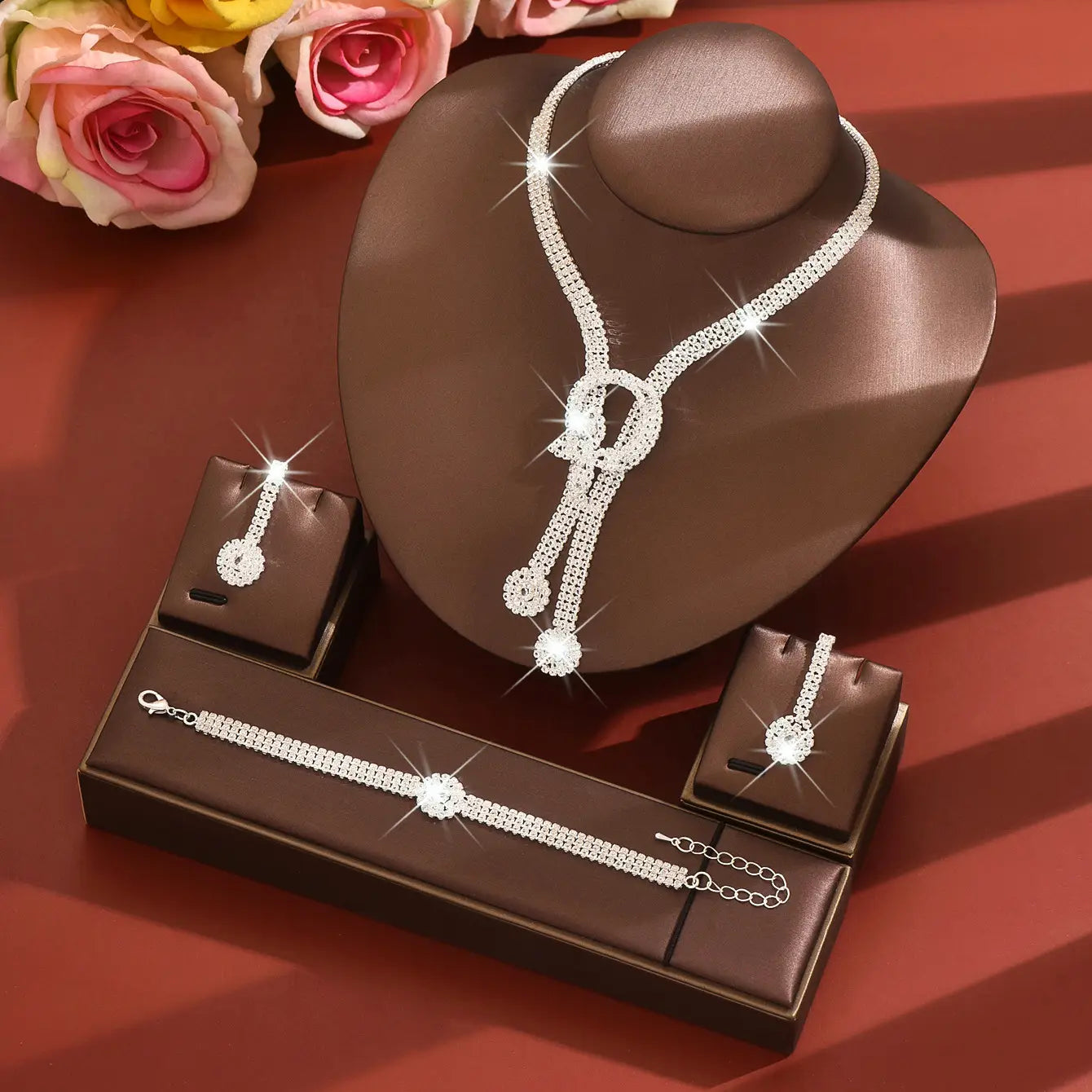Parure de bijoux de mariage en diamant argentée et scintillante, avec un collier en cascade orné de cristaux transparents et d’un motif central de nœud, présenté sur un buste de mannequin brun, accompagnée de boucles d’oreilles et d’un bracelet assortis.