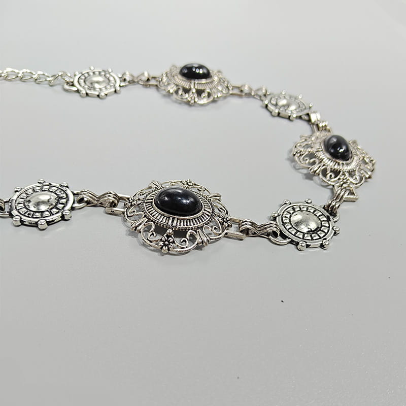 Ceinture de hanche en chaîne pour cosplay argentée, comportant des maillons de style vintage ornés avec des pierres ovales noires serties dans un travail métallique en filigrane complexe, pour un style gothique punk.