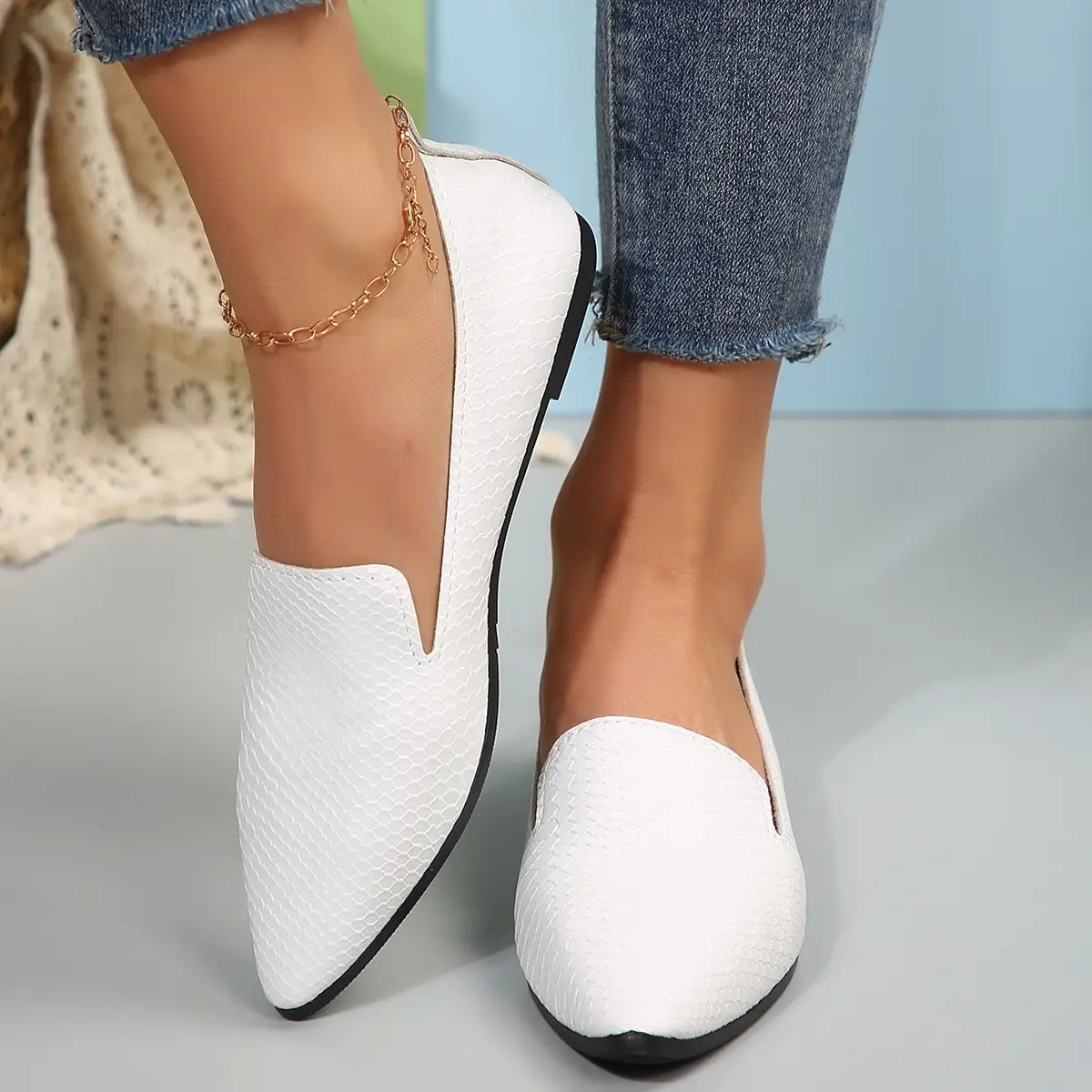Mocasines planos de cuero con punta afilada para poner y quitar con facilidad, con textura de piel de serpiente blanca, suelas negras y pulsera tipo gargantilla dorada para el tobillo.