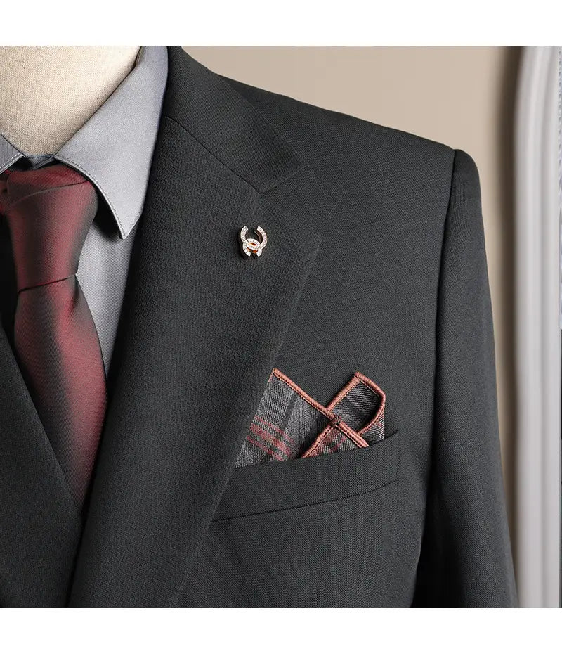 Petite épingle à tonalité argentée avec un motif de corne stylisé et des accents rouges, fixée au revers d’un ensemble costume d’affaires pour homme gris foncé slim fit en trois pièces, adapté aux soirées décontractées et aux occasions d’affaires en mode masculine.