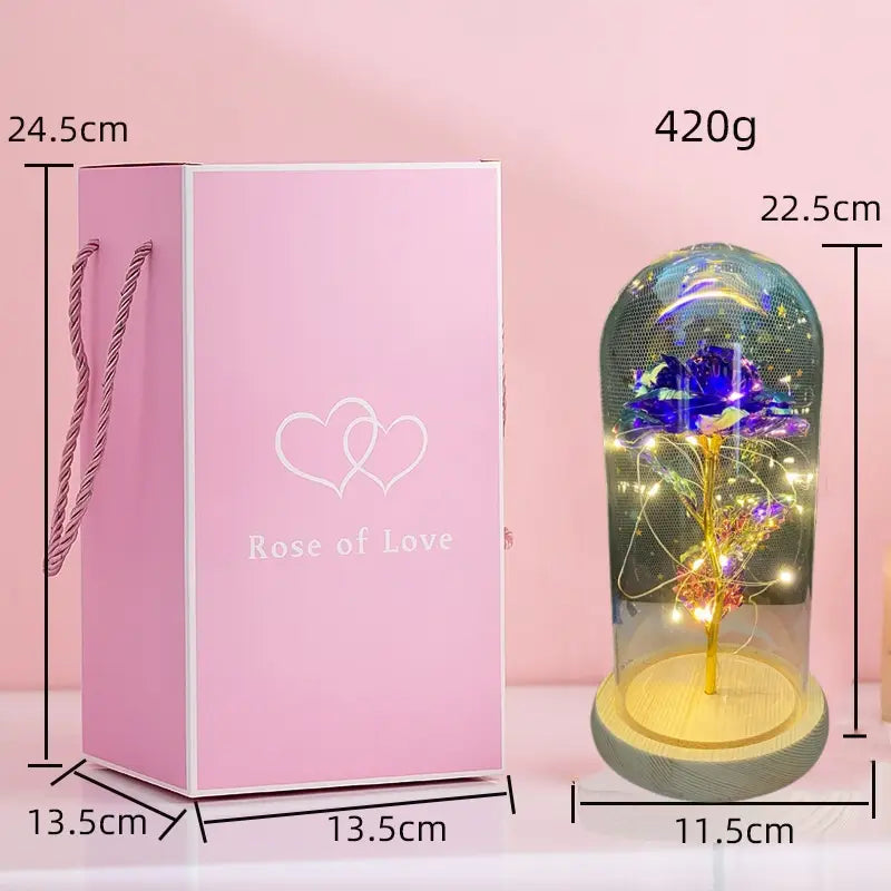 Eine leuchtend blaue und violette konservierte Rose mit goldenem Stiel, umschlossen in einer klaren Glaskuppel, auf einem Holzsockel, mit LED-Licht im „Eternal Gold Rose Glass Dome Gift“.