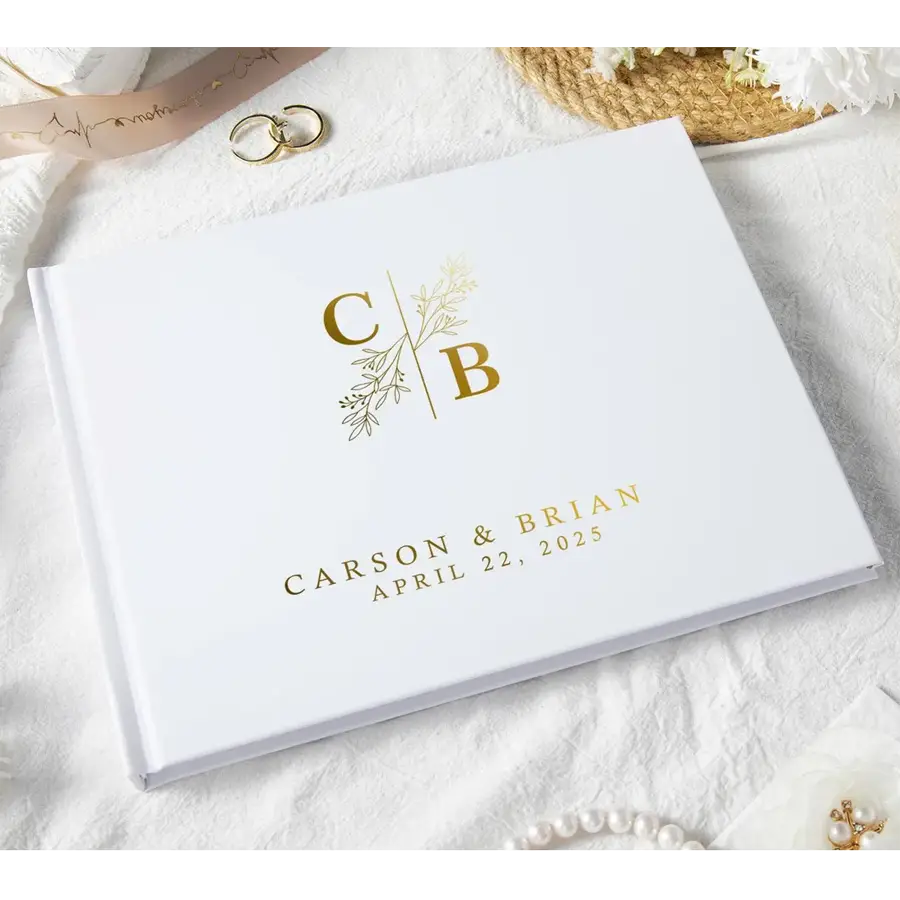 個別にパーソナライズされたブラック＆ホワイトの結婚ゲストブック記念品。氏名「Carson」と「Brian」、日付「4月22日」を記載し、繊細なボタニカルデザインで区切られたイニシャル「c」と「b」を、ゴールド箔の刻印で表したホワイトカバーを特徴とします。