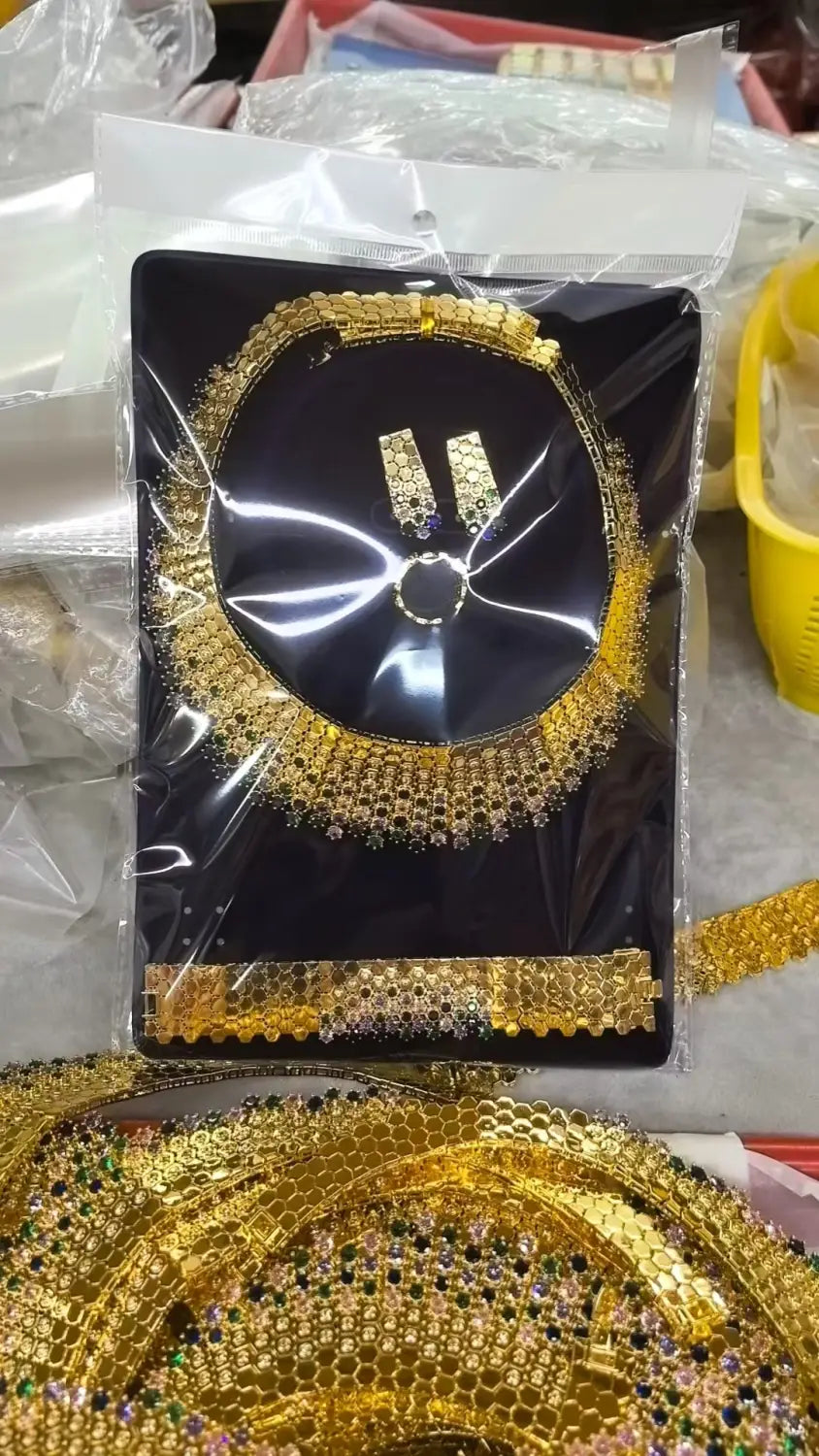 Ensemble de bijoux géométriques en zircon doré comportant un collier de couleur dorée, avec des détails complexes de perlage et de pierres précieuses, présentés sur une carte noire dans un emballage en plastique transparent.