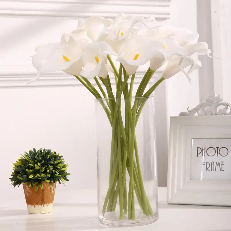 a bouquet of artificial white calla lilies with yellow centers arranged in a clear glass vase for home decor.