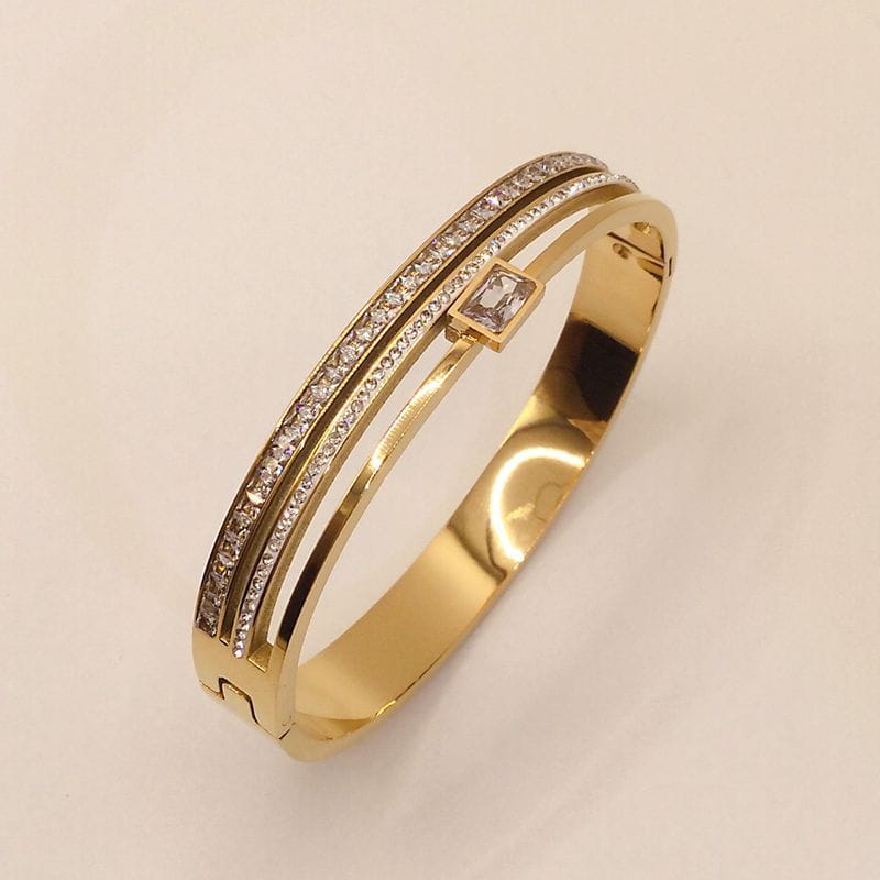 Armreif aus rostfreiem Stahl in Goldtönung mit Kristallen, mit zwei parallelen Bändern, einer Reihe kleiner Diamanten und einem größeren quadratisch geschliffenen Diamant-Akzent.