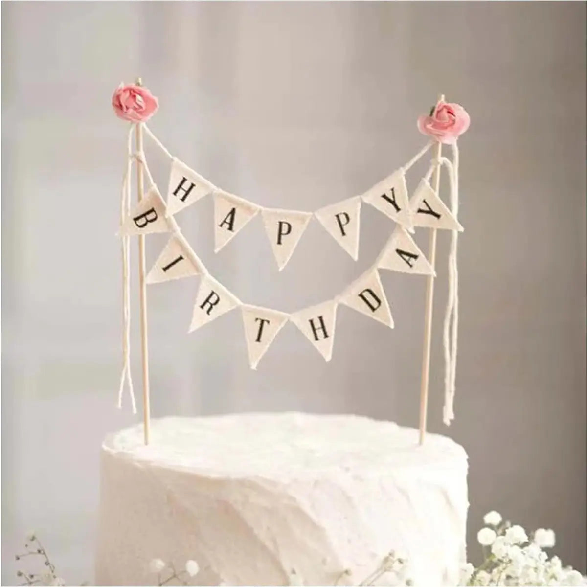 Un délicat fanion de gâteau d’anniversaire couleur crème, surmonté d’une inscription noire et de roses en tissu roses à chaque extrémité, repose sur un gâteau blanc glacé.