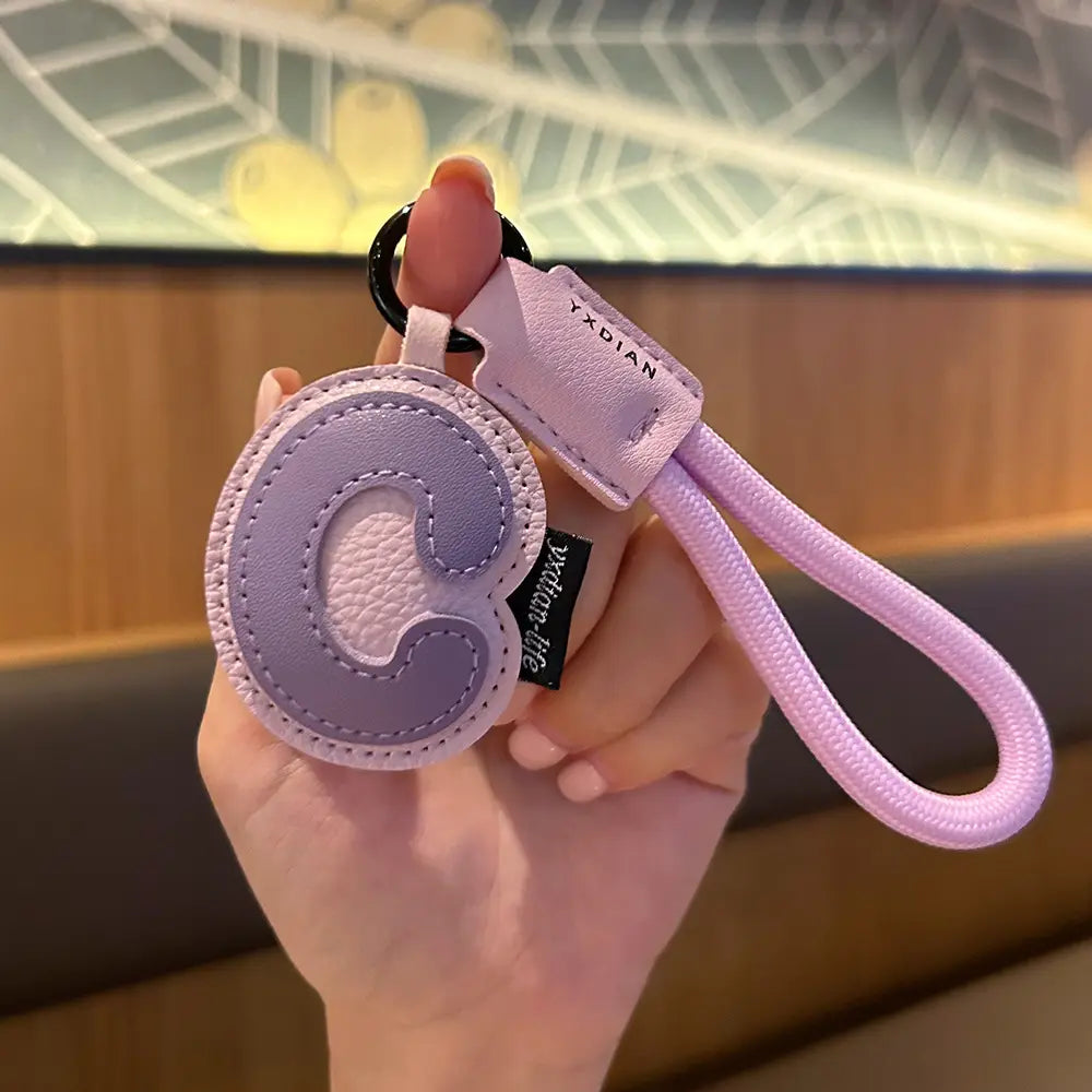 A lavender leather initial keychain wristband charm featuring a circular letter c design with contrasting stitching and a matching fabric loop.