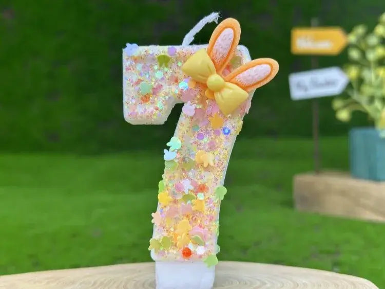 A glitter bunny ear number birthday candle featuring a glittery pastel-colored number seven decorated with a yellow bow and bunny ears.