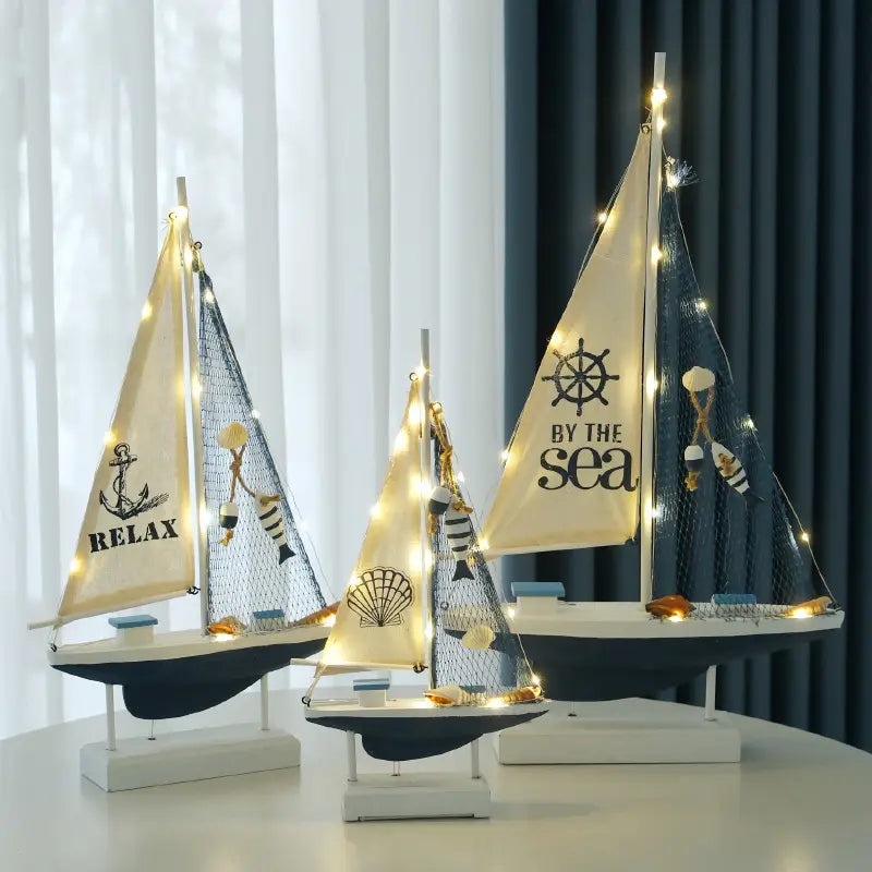 Drei Modelle einer LED-Segelboot-Dekoration im mediterranen Stil aus Holz für die Wohnraumgestaltung: beleuchtete Segel in Creme und Navyblau mit Netzdetails sowie aufgedruckten maritimen Motiven wie Anker und Schiffsräder.