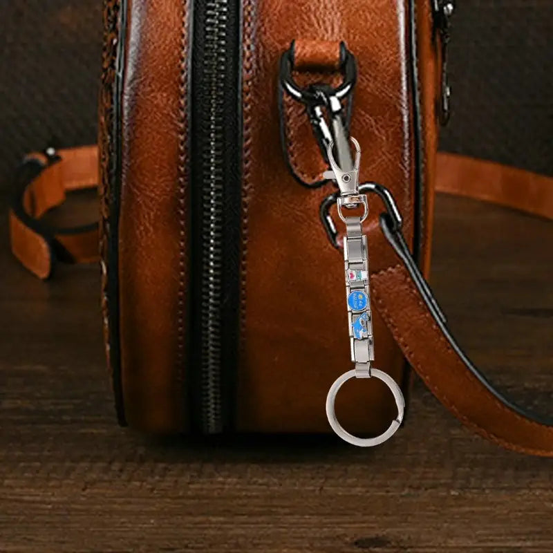 Un llavero con dije de eslabones italianos con acabado plateado, con un dije rectangular esmaltado azul y blanco sujeto a un bolso de cuero marrón mediante un broche de metal negro.