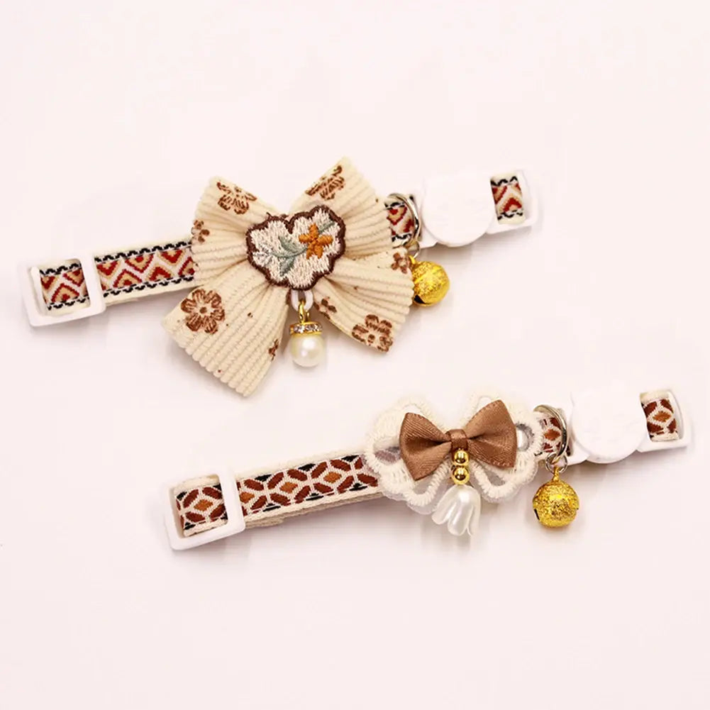 adjustable floral cat collars featuring a cream-colored ribbed bow with embroidered floral details and pearl pendant, and a brown satin bow with lace trim and gold bell charms.